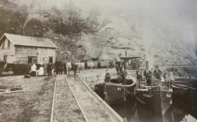 FRA TOG TIL BÅT. Hellarmo stasjon 1895. Her var det omlasting fra jernbane til båt. D/S «Sulitelma I» med passasjerer og lastede prammer var klar for opp-transport. Dampkranen ble brukt ved lossing av kis fra pram til jernbanevogn. FRA TOG TIL BÅT. Hellarmo stasjon 1895. Her var det omlasting fra jernbane til båt. D/S «Sulitelma I» med passasjerer og lastede prammer var klar for opp-transport. Dampkranen ble brukt ved lossing av kis fra pram til jernbanevogn. Foto: Sulitjelma Gruber AS