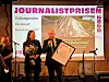 To personer på scene mottar journalistpris med diplom og blomster foran stor skjerm.