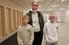 Tre personer står i en lys innendørs hall med tredører og skytebane i bakgrunnen.