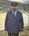 En mann i mørk uniform og hatt står ute ved vann med fjell i bakgrunnen.