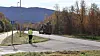 Person ved veikryss med høsttrær, fjell og grønt kjøretøy i bakgrunnen.